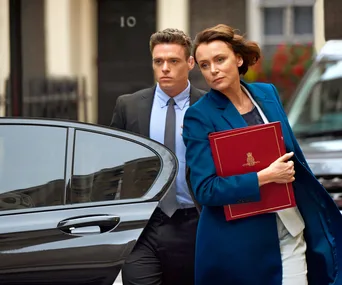 A woman in a blue coat holds a red folder, followed by a man in a suit exiting a car outside a building.