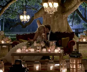 Couple kissing on a couch under a tree, surrounded by candles and chandeliers, creating a romantic ambiance.