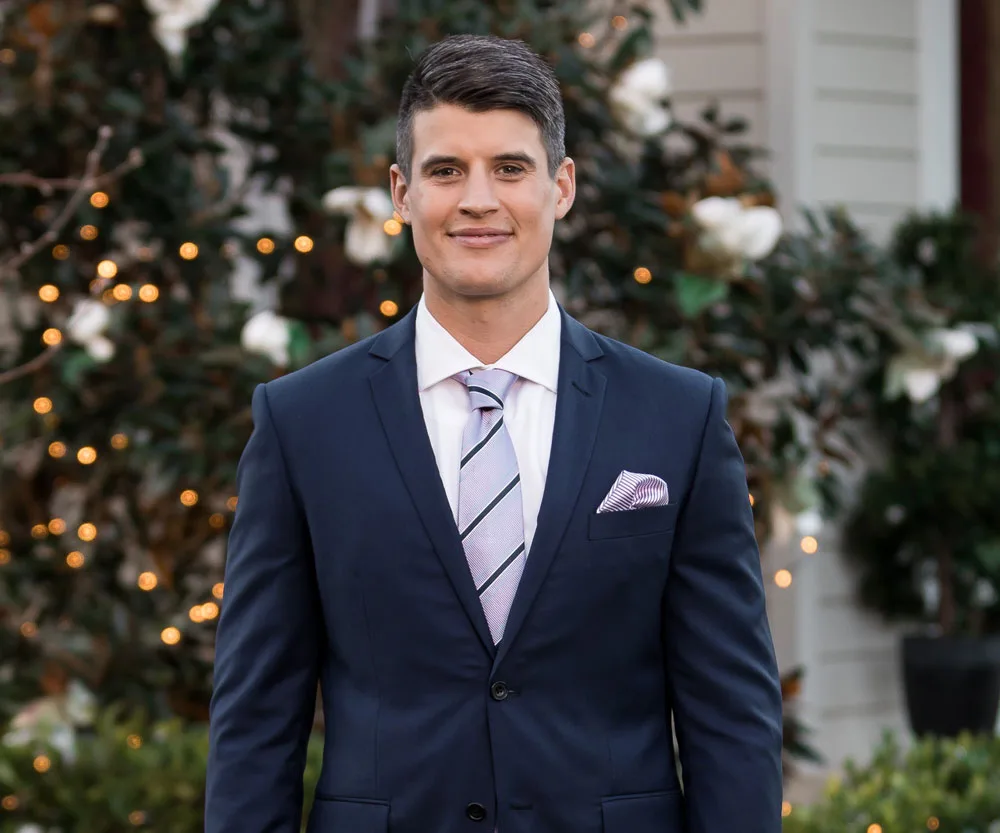 Man in a blue suit with a pink tie and pocket square, standing outdoors with greenery and lights in the background.