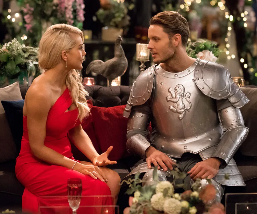 A woman in a red dress talks to a man in knight armor on a sofa, surrounded by plants and decorative lights.