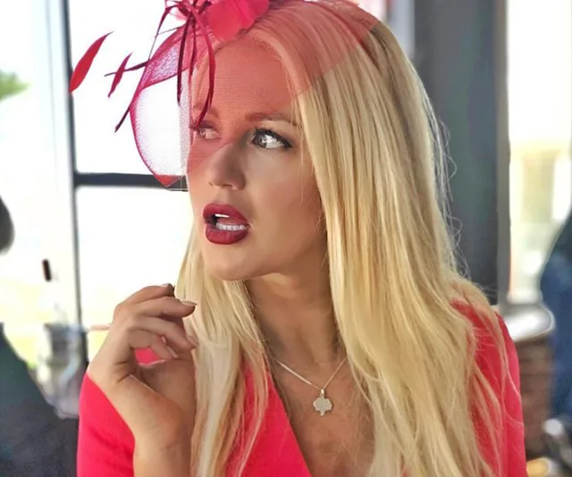 Blonde woman in a red outfit with a fascinator, looking contemplatively to the side.