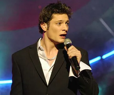 Young man in a suit singing passionately on stage with a microphone, blue lights in the background.