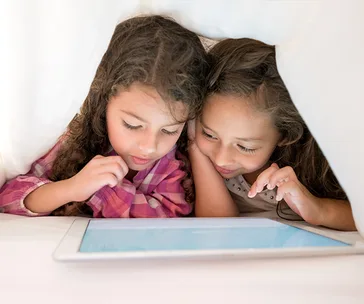 Two children under a blanket intently focused on a tablet screen.