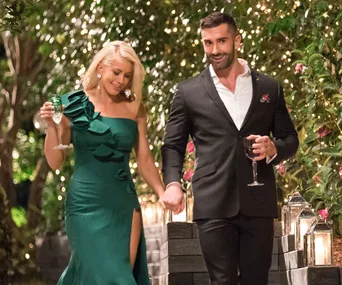 A couple holding hands and smiling, walking through a garden adorned with lanterns and fairy lights, dressed elegantly.