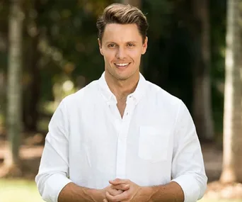 Man in a white shirt smiling outdoors with trees in the background.