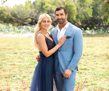 A couple poses outdoors; the woman in a navy dress and the man in a light blue suit, smiling at the camera.