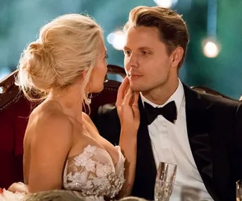 A couple in formal attire share an intimate moment, holding champagne glasses, under warm outdoor lighting.