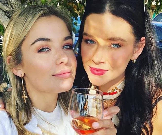 Two women smiling, one holding a wine glass, outdoors with sunlight filtering through trees.