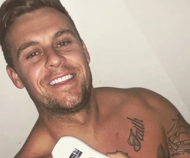 A shirtless man with tattoos and a smile, holding a remote control against a plain background.