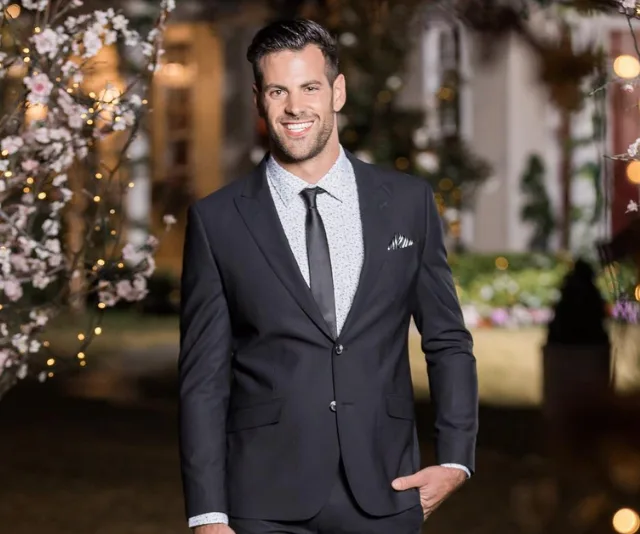 A man in a navy suit stands smiling outdoors, surrounded by lights and flowering trees.