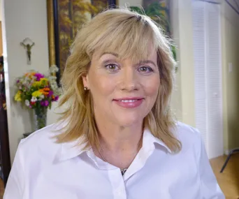 A woman with blonde hair wearing a white shirt smiles while sitting indoors with flowers in the background.
