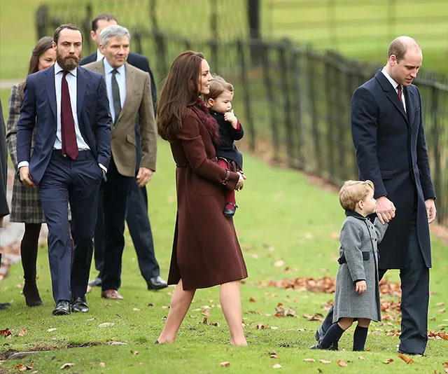 Royal family, including a couple with children, walk outdoors on grass, accompanied by others in formal clothing.