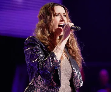 Singer performing passionately on stage with a microphone, wearing a patterned jacket and sparkling dress.