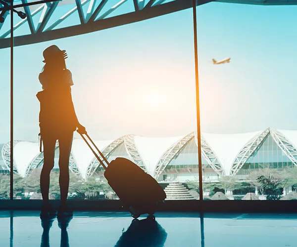 Silhouette of a traveler with a suitcase, looking at a flying airplane through large airport windows at sunset.