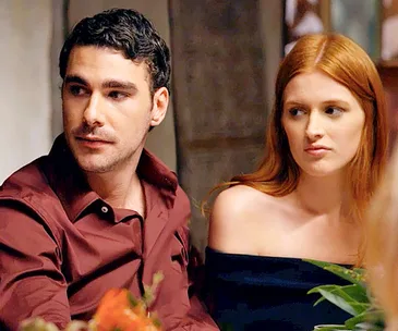 Two people sitting at a table; the man in a maroon shirt, and the woman with red hair, looking intently at something.