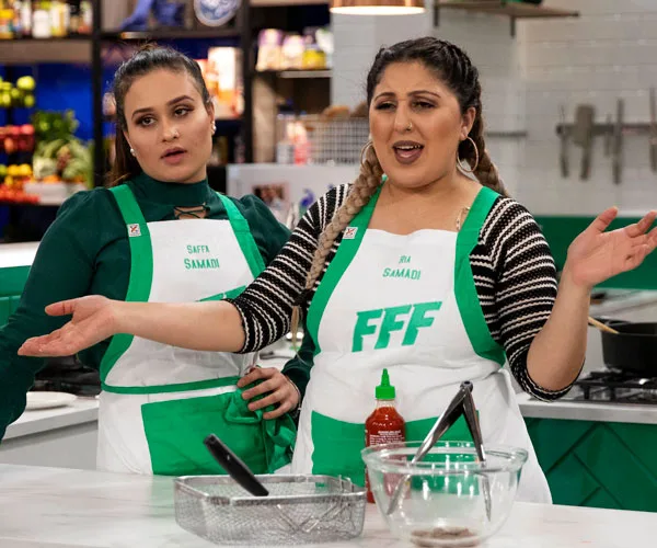 Samadi sisters in kitchen during Family Food Fight, wearing aprons, engaged in a lively cooking discussion.