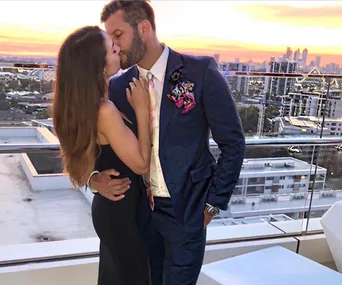 A couple embraces and kisses on a rooftop at sunset, overlooking a cityscape.