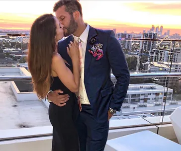 A couple embraces and kisses on a rooftop at sunset, overlooking a cityscape.