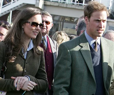 A couple walks outdoors wearing formal attire; the woman has sunglasses, and the man wears a suit, among a crowd.