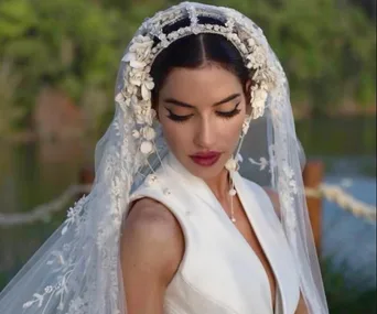 A bride in a white dress and floral veil, looking down with a serene expression, standing outdoors.