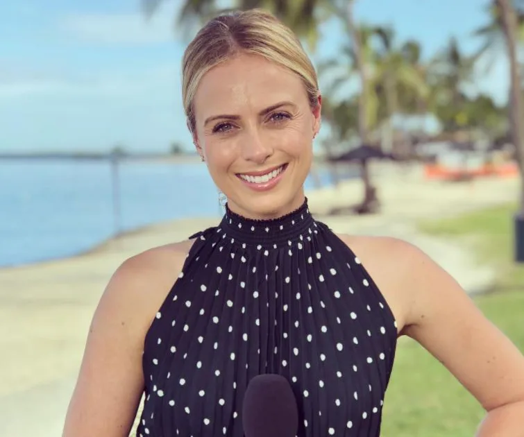 A woman in a polka dot dress smiles while holding a microphone on a beach with palm trees and the sea in the background.