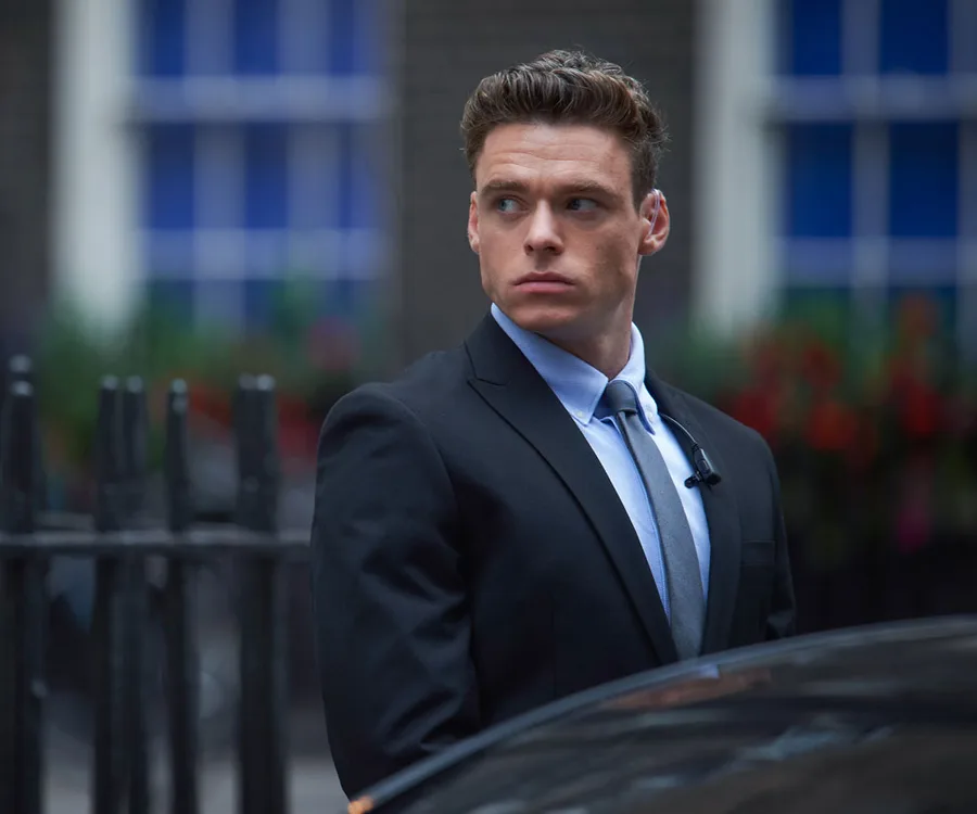 David Budd from "Bodyguard" in a suit, looking to the side, with a building and railing in the background.