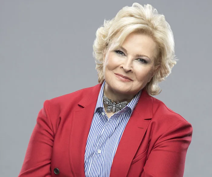 A woman with short blonde hair in a red blazer and striped shirt smiles confidently against a gray background.