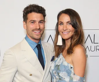 Two smiling individuals pose together; one in a light suit and the other in a floral dress with statement earrings.