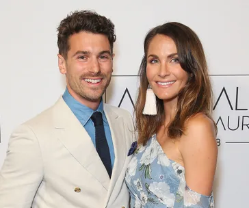 Two smiling individuals pose together; one in a light suit and the other in a floral dress with statement earrings.