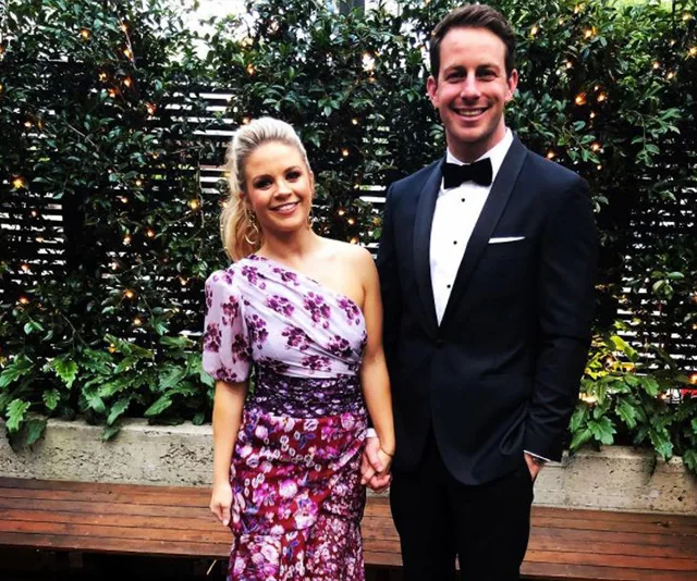 A couple dressed formally, posing outdoors in front of greenery.