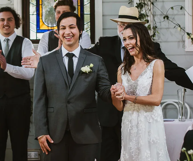 Bride and groom smiling, holding hands at a wedding ceremony, with guests applauding in the background.