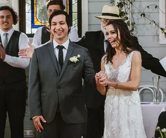 Bride and groom smiling, holding hands at a wedding ceremony, with guests applauding in the background.