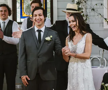 Bride and groom smiling, holding hands at a wedding ceremony, with guests applauding in the background.