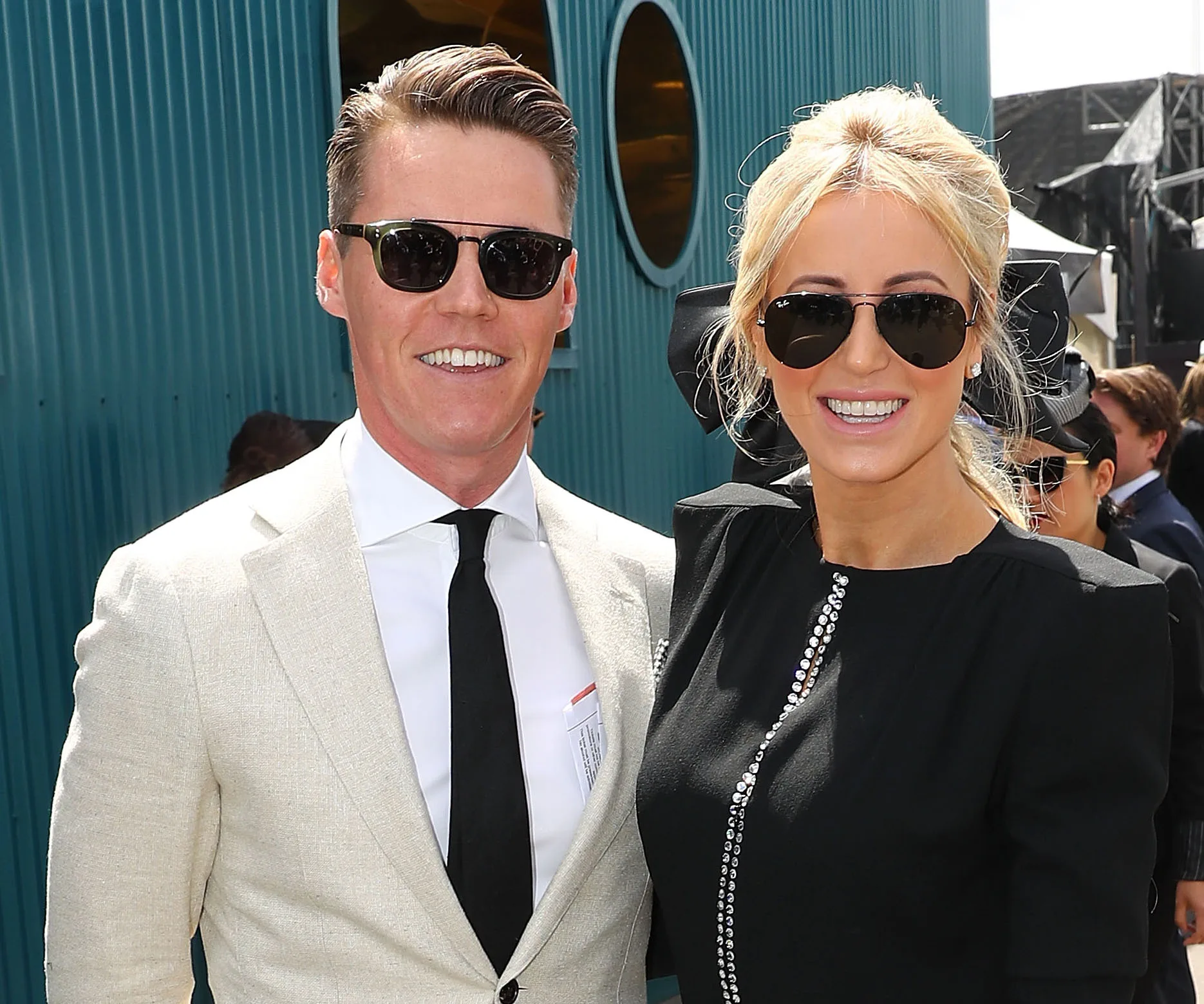 Two people smiling in sunglasses at an outdoor event, one in a black outfit, the other in a light suit and tie.