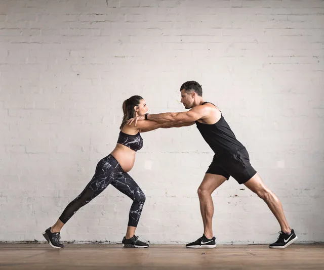 Pregnant woman and man exercising together, performing a partner lunge in workout gear.