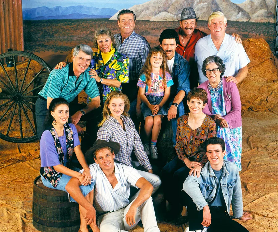 Cast of "A Country Practice" in a group photo with countryside backdrop, featuring actors in casual attire.