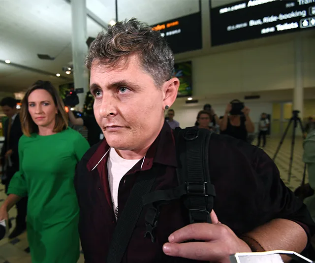 A person with short hair at an airport, holding a backpack; surrounded by people and photographers.