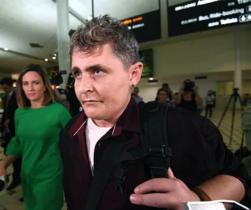 A person with short hair at an airport, holding a backpack; surrounded by people and photographers.