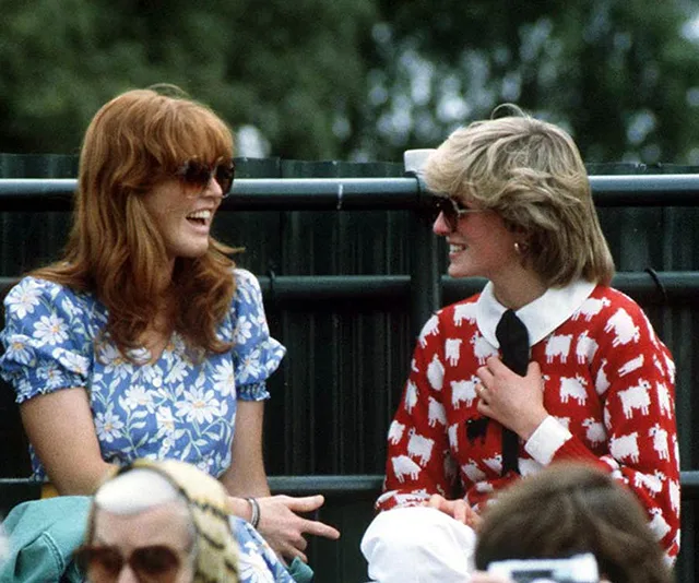 Two women, one in a blue floral dress and the other in a red sheep-patterned sweater, laughing outdoors in sunglasses.