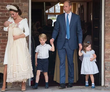 A family of five stands together, including a baby in a christening gown, outdoors near a brick building.