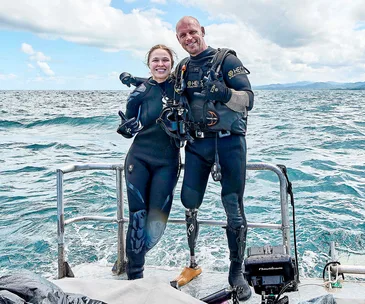 Two divers in wetsuits smiling on a boat with ocean backdrop, one has prosthetic legs.