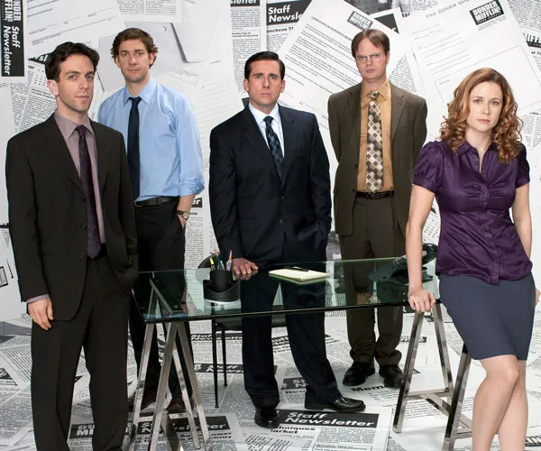 A group of five office employees standing around a desk, in a room with paper-covered walls and floor.