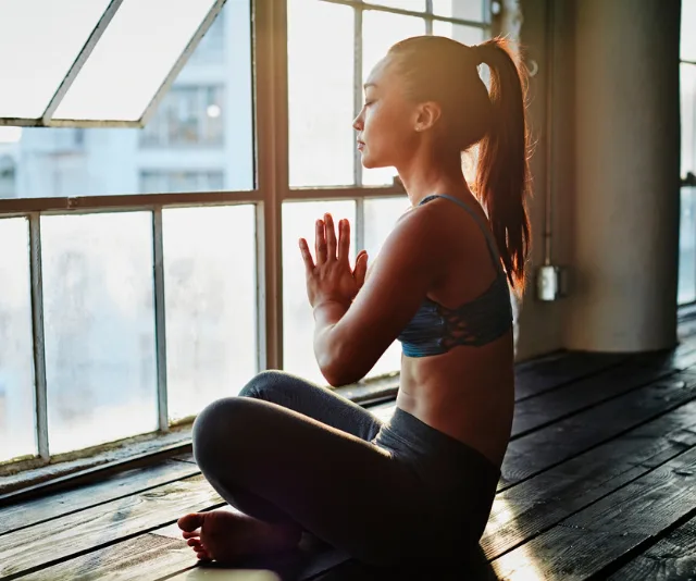 A woman practicing meditation, sitting cross-legged by a large window, with hands in a prayer position, wearing athletic wear.