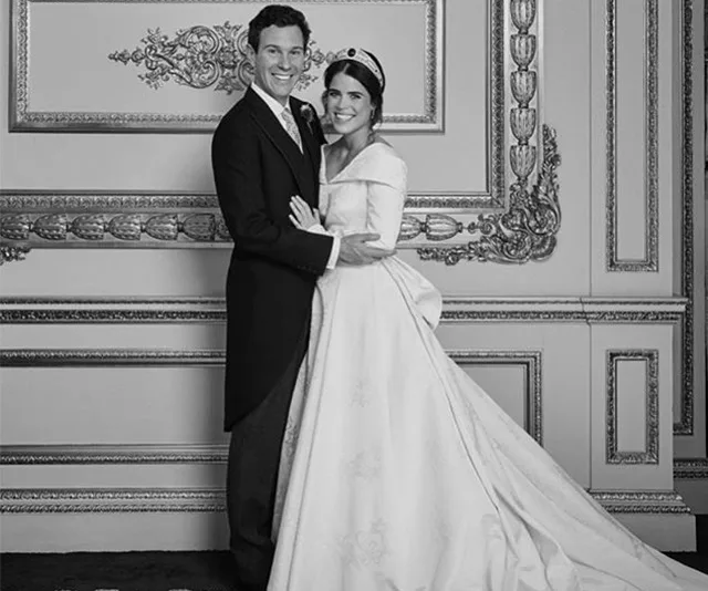 A bride in a gown and groom in a suit pose together in an ornate room, both smiling warmly.
