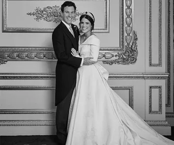 A bride in a gown and groom in a suit pose together in an ornate room, both smiling warmly.