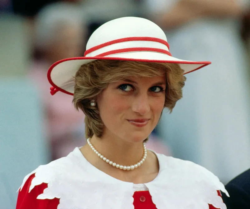 A woman in a white and red hat with pearls, smiling subtly.