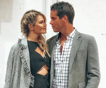 A man and woman stand closely, gazing at each other with smiles, dressed in formal attire against a plain backdrop.