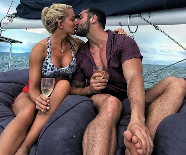A couple kissing on a sailboat, holding glasses of white wine, against an ocean backdrop.