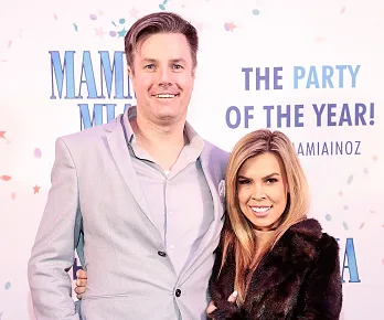 A couple poses at the "Mamma Mia! The Party" event backdrop, smiling, with celebratory text and confetti graphics in the background.