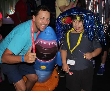 Man posing with inflatable shark and a child with a shiny wig and party accessories at a colorful event.